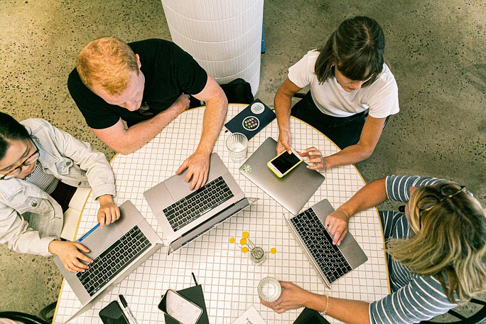 A group of people sitting around a table with laptops, engaged in discussion. The setting is indoors, with a wall visible in the background. The participants include men and women, each focused on their computers.